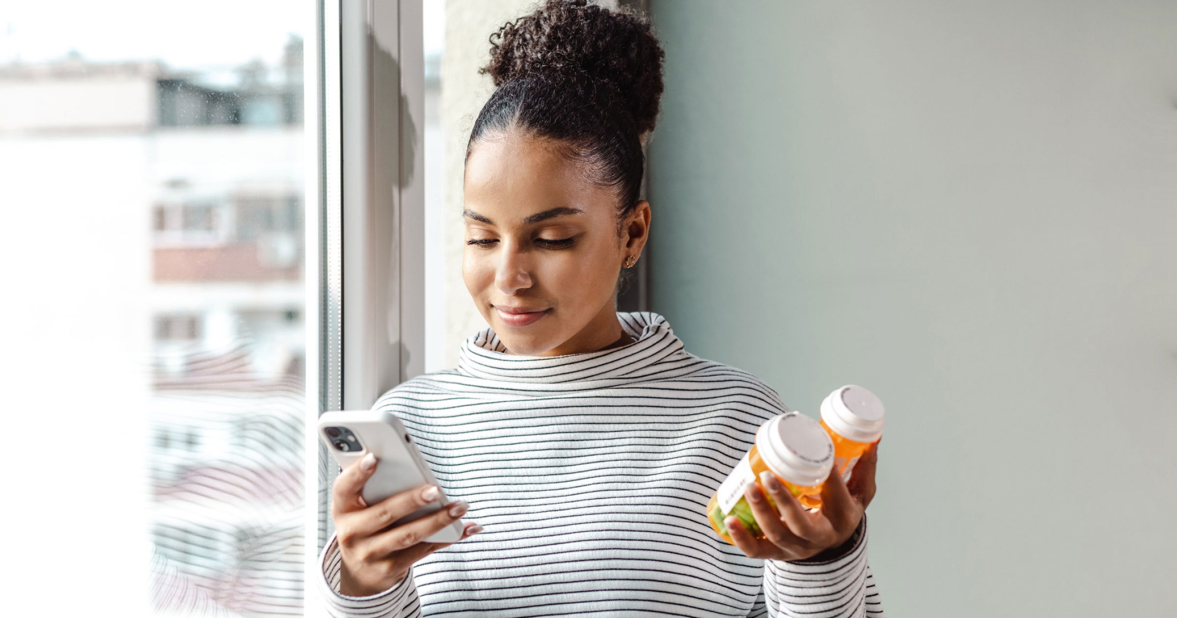 A photo of a woman holding pill bottles in one hand while ordering medication online by using her phone.