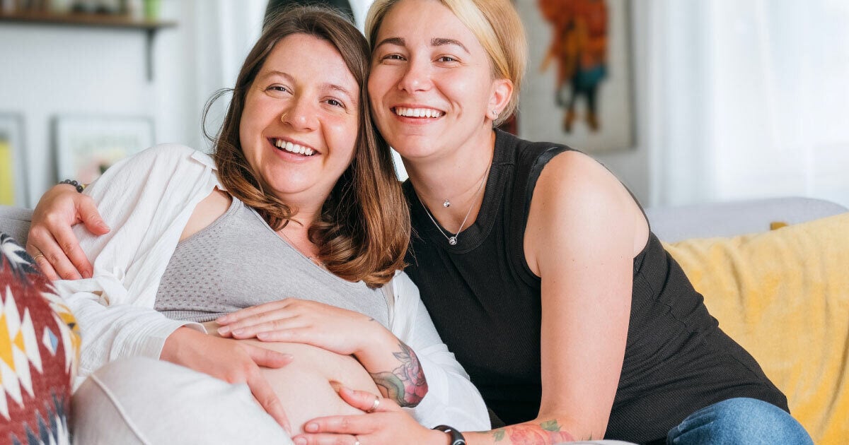 A same sex couple smile at the camera. One partner is pregnant.