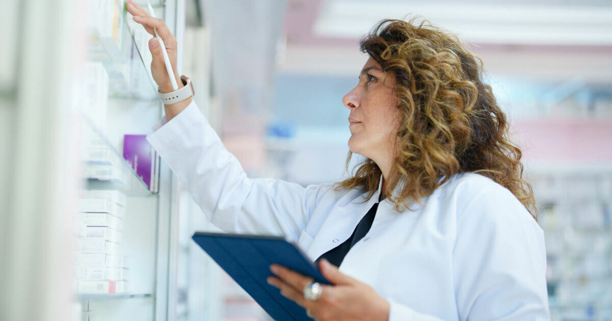 A woman in a lab coat is standing in front of shelving and is holding a tablet while looking for a specific prescription.